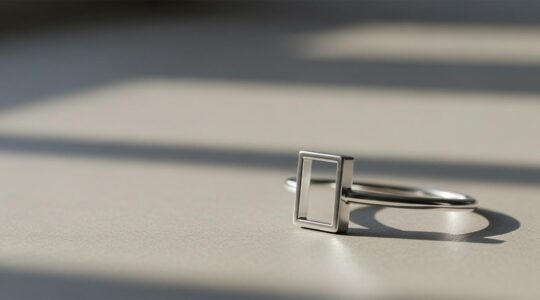 Un bijou minimaliste élégant, lumineux, reposant sur un fond neutre avec un jeu subtil d'ombres et lumière mettant en valeur la simplicité et la force du design épuré